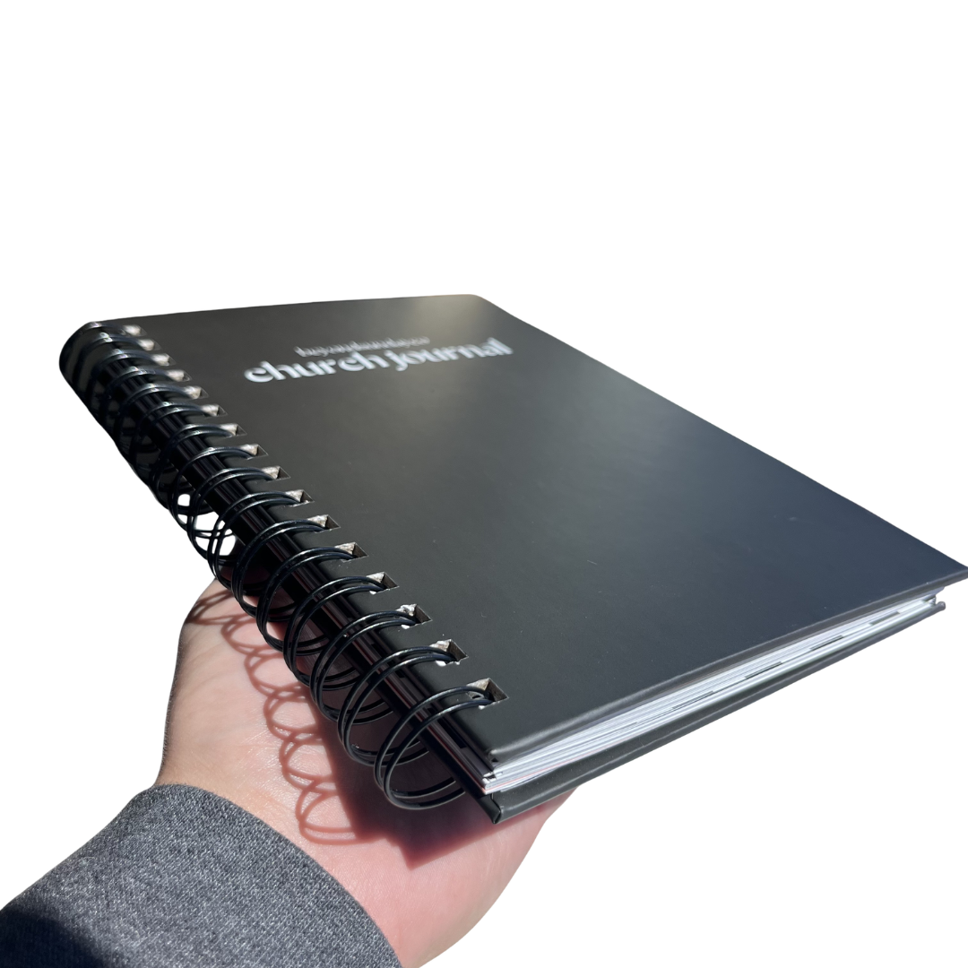 Black spiral-bound notebook titled 'church journal' held in a hand on a white background